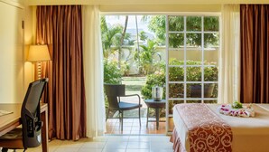 A hotel room with a bed, a desk, a chair, and a view of a garden through a glass door. - Bay Gardens Hotel (Gros Islet)