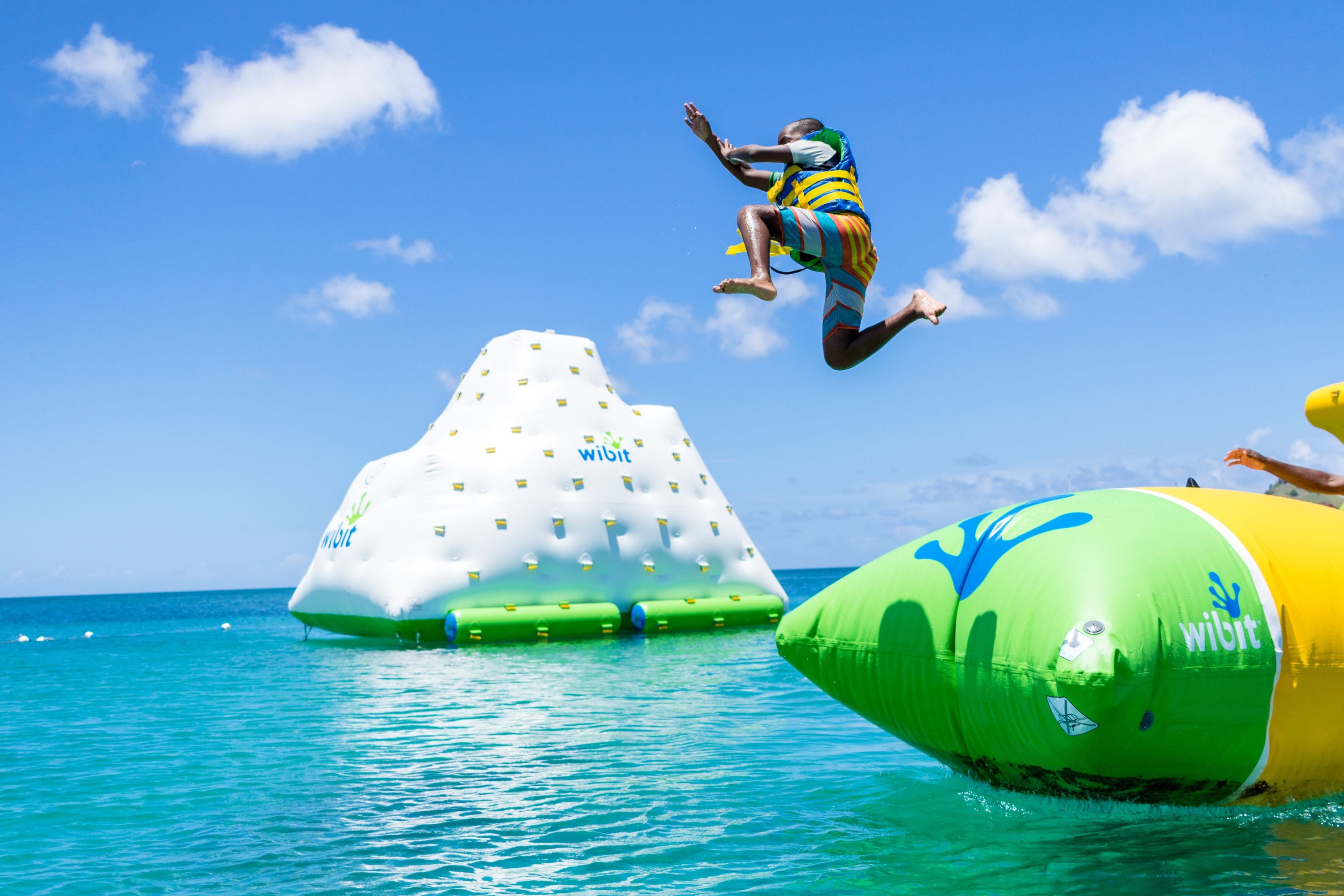 Una persona salta desde una estructura inflable verde y amarilla hacia el agua, con una isla inflable blanca con la marca "Wibit" al fondo.