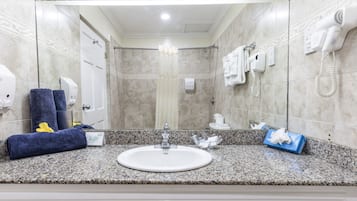 A bathroom with a granite countertop, a white sink, a mirror, a towel rack with towels, a hairdryer, and a shower area.