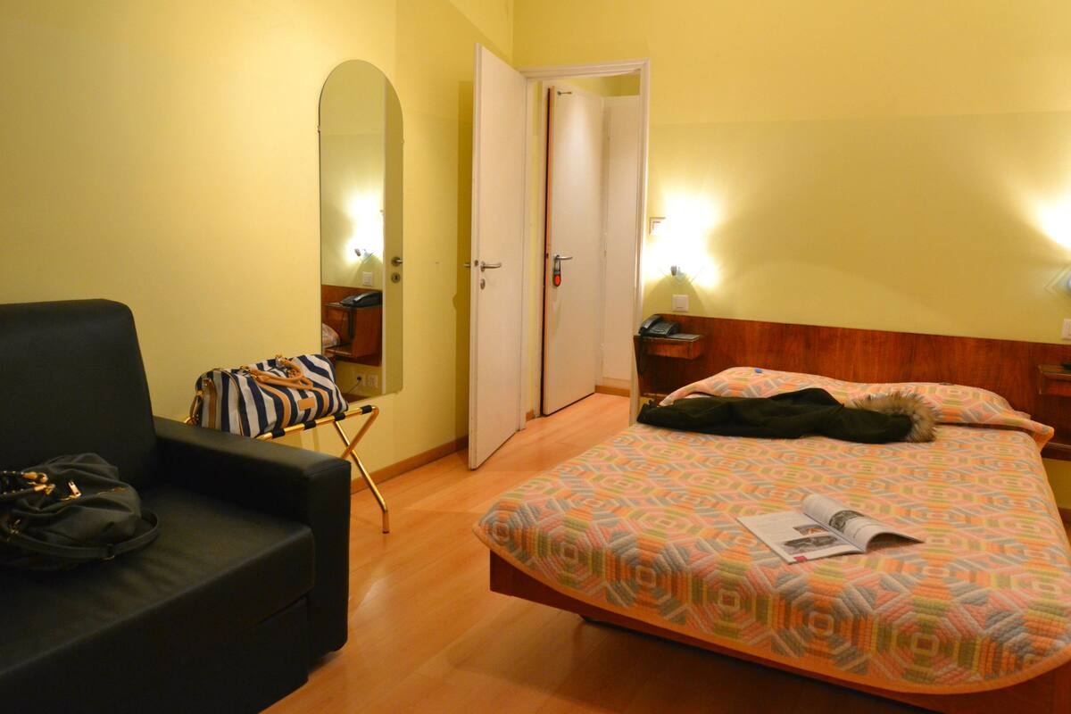Una habitación de hotel con cama, una silla, mesita de noche, espejo y puerta.