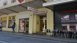 Entrada de un hotel con carteles que indican 'HOTEL MODERNE' en francés. Hay mesas y sillas al aire libre, y una persona caminando cerca.