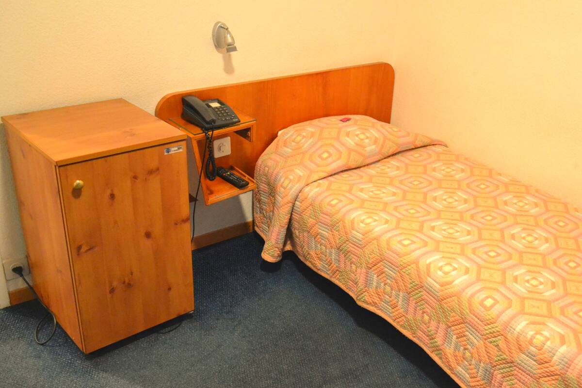 Una habitación pequeña con cama individual, cabecera de madera, mesita de noche con teléfono y un mueble auxiliar.