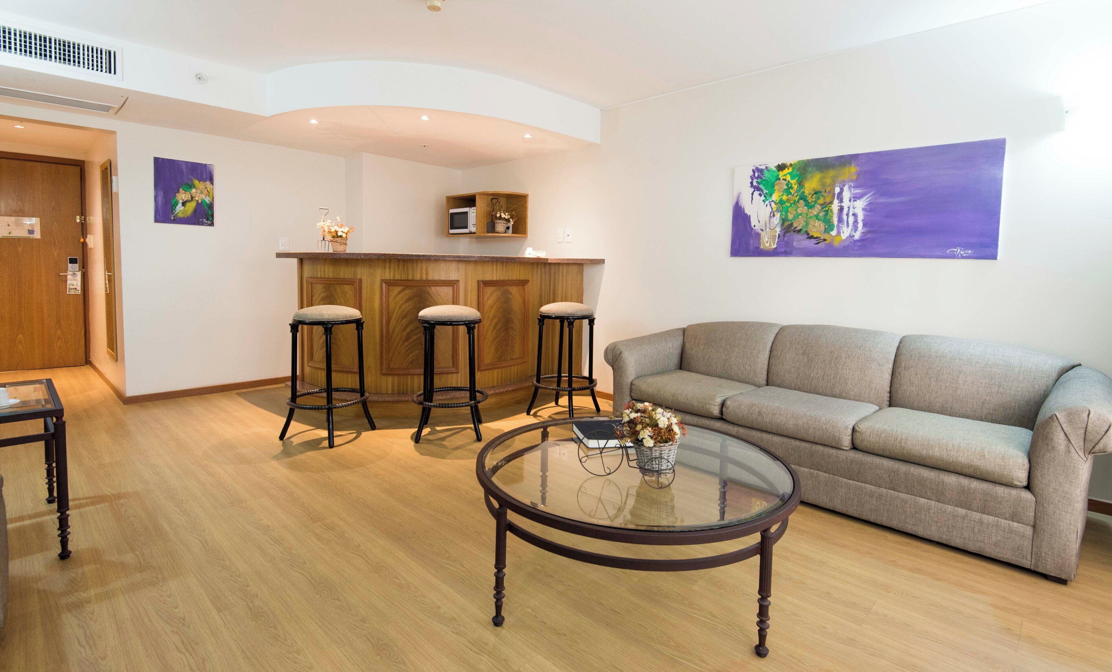 Uma sala de estar moderna com uma mesa de centro de vidro, um sofá cinza e três banquetas.