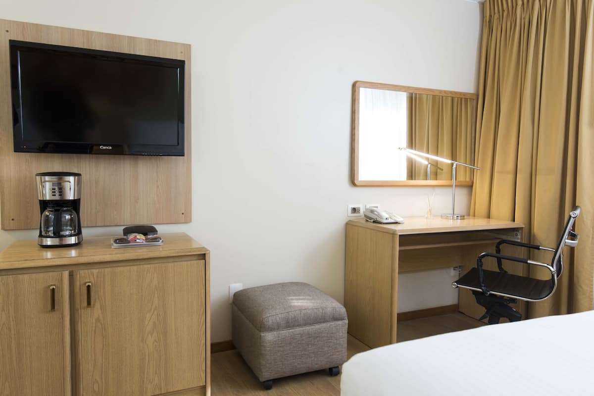 A hotel room with a flat-screen TV, a coffee maker, a desk with a chair, a small ottoman, and a mirror.