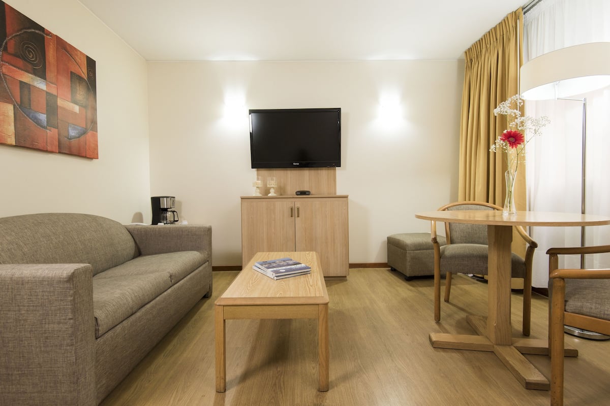 A modern hotel room with a sofa, a wooden coffee table, a TV mounted on a cabinet, and a dining area with a table and chairs.