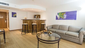 A living room with a grey sofa, a round glass coffee table, and three bar stools. A kitchen area with wooden cabinets and a microwave is visible in the background. - Holiday Inn Montevideo by IHG (Montevideo)