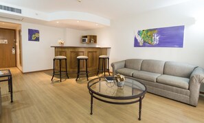 Un salon avec un canapé gris, une table basse ronde en verre et trois tabourets de bar. On aperçoit à l’arrière-plan un coin cuisine avec des meubles en bois et un four à micro-ondes.