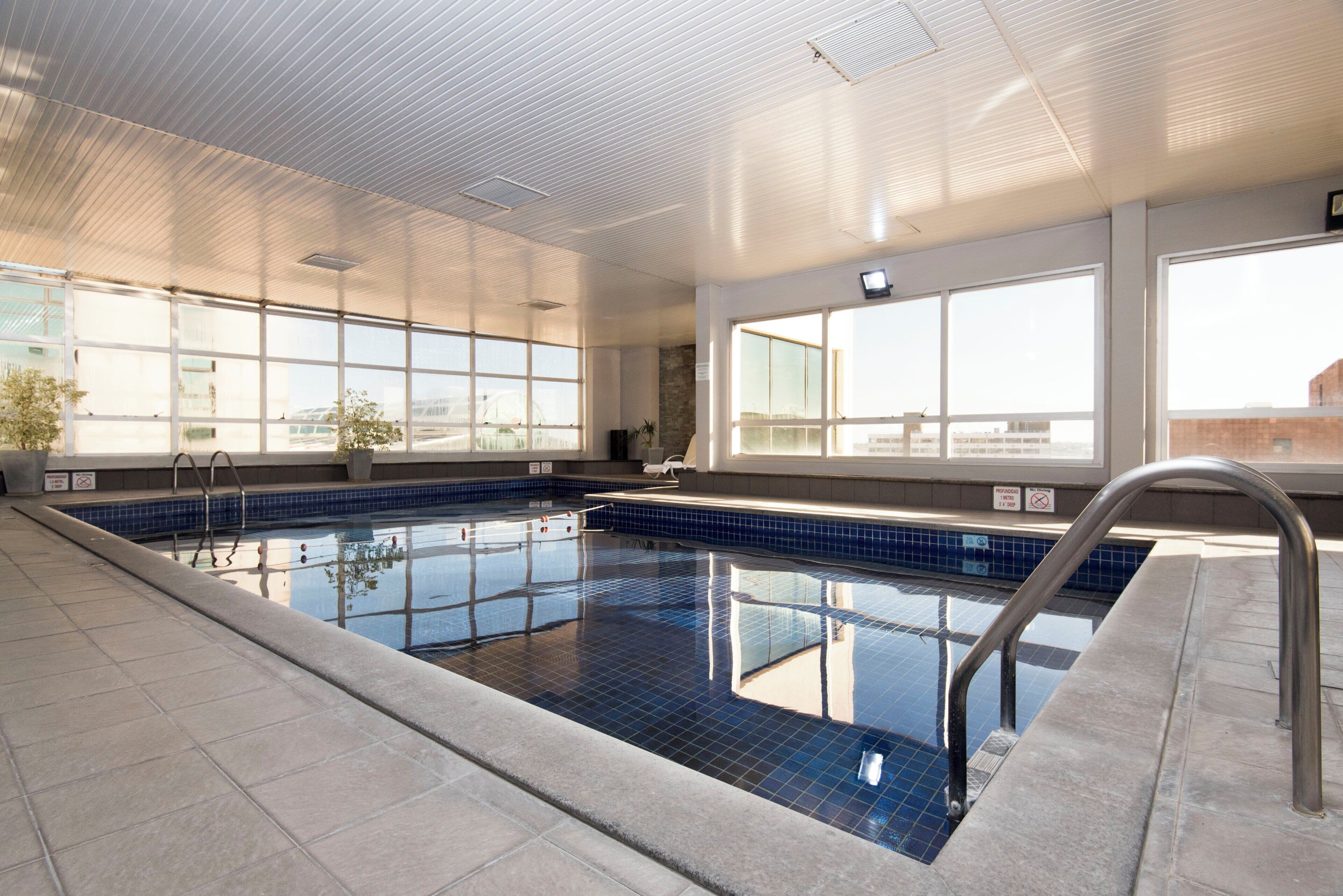 An indoor swimming pool with clear blue tiles, a metal ladder, and large windows allowing natural light.
