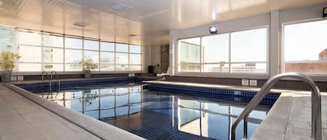 Una piscina interior con baldosas azules cristalinas, una escalera metálica y grandes ventanales que permiten la entrada de luz natural.