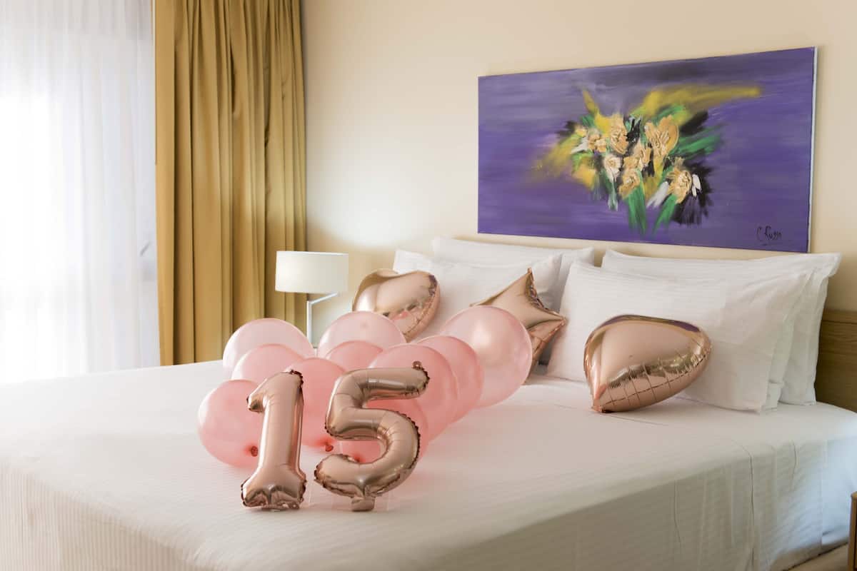 A hotel room with a bed, white bedding, a painting on the wall, and a bouquet of balloons.
