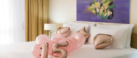 A hotel room with a bed, white bedding, a painting on the wall, and a bouquet of balloons.