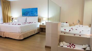 A hotel room with a double bed, a side table, a nightstand, a large mirror, a painting on the wall, and a bathtub with rose petals.