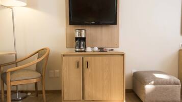 Una habitación con un armario de madera, un televisor montado encima, una cafetera, un taburete acolchado y una silla con cojín.