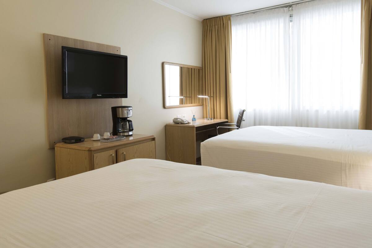 A hotel room with two beds, a desk, a coffee maker, a TV, and a window with curtains.