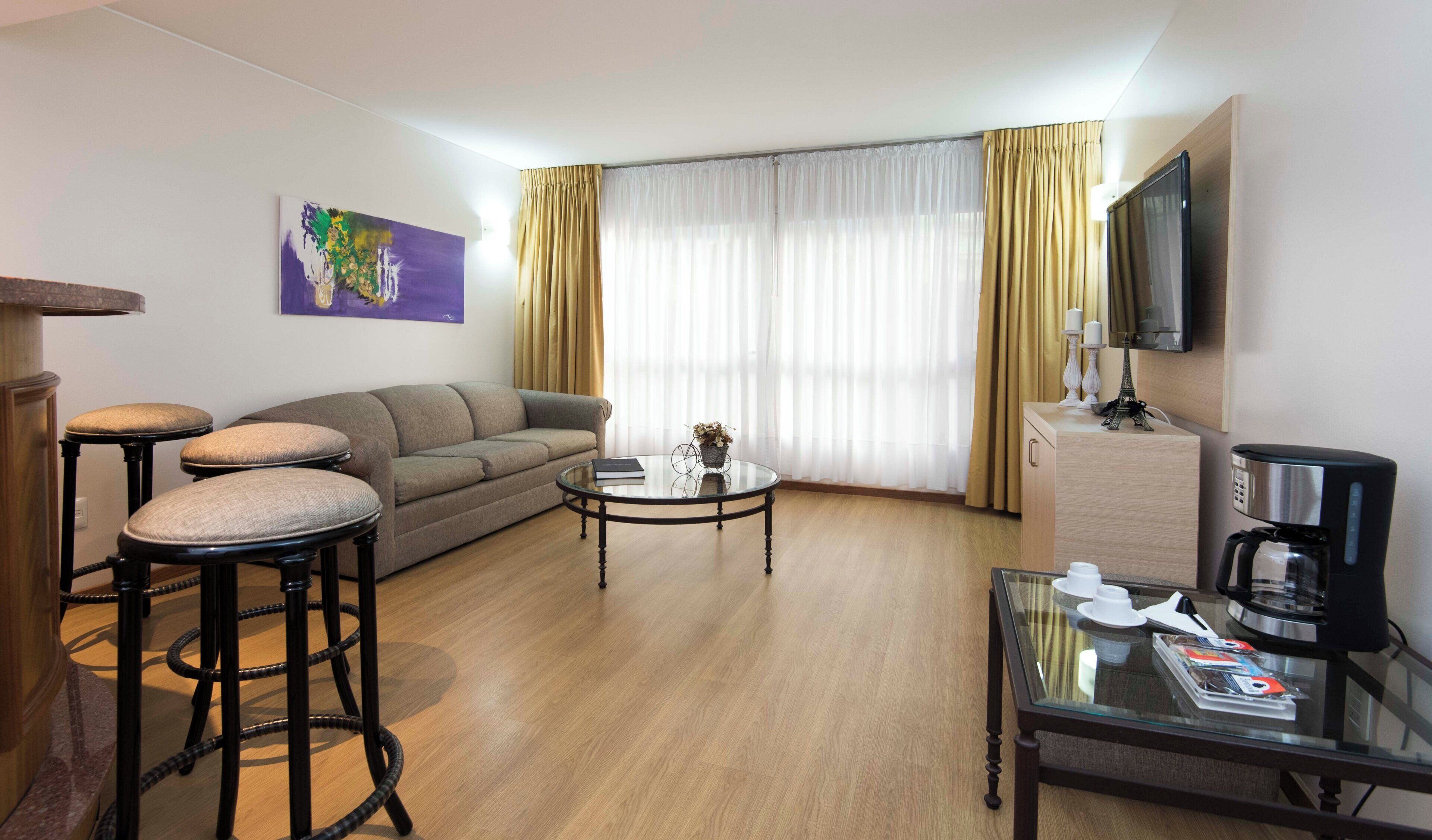 A hotel room with a sofa, two bar stools, a coffee table, a television, and a minibar.