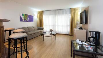 A hotel room with a sofa, two bar stools, a coffee table, a television, and a minibar.