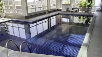 Una piscina interior con baldosas azules cristalinas, una escalera de metal y una zona de estar con sillas y una mesa.