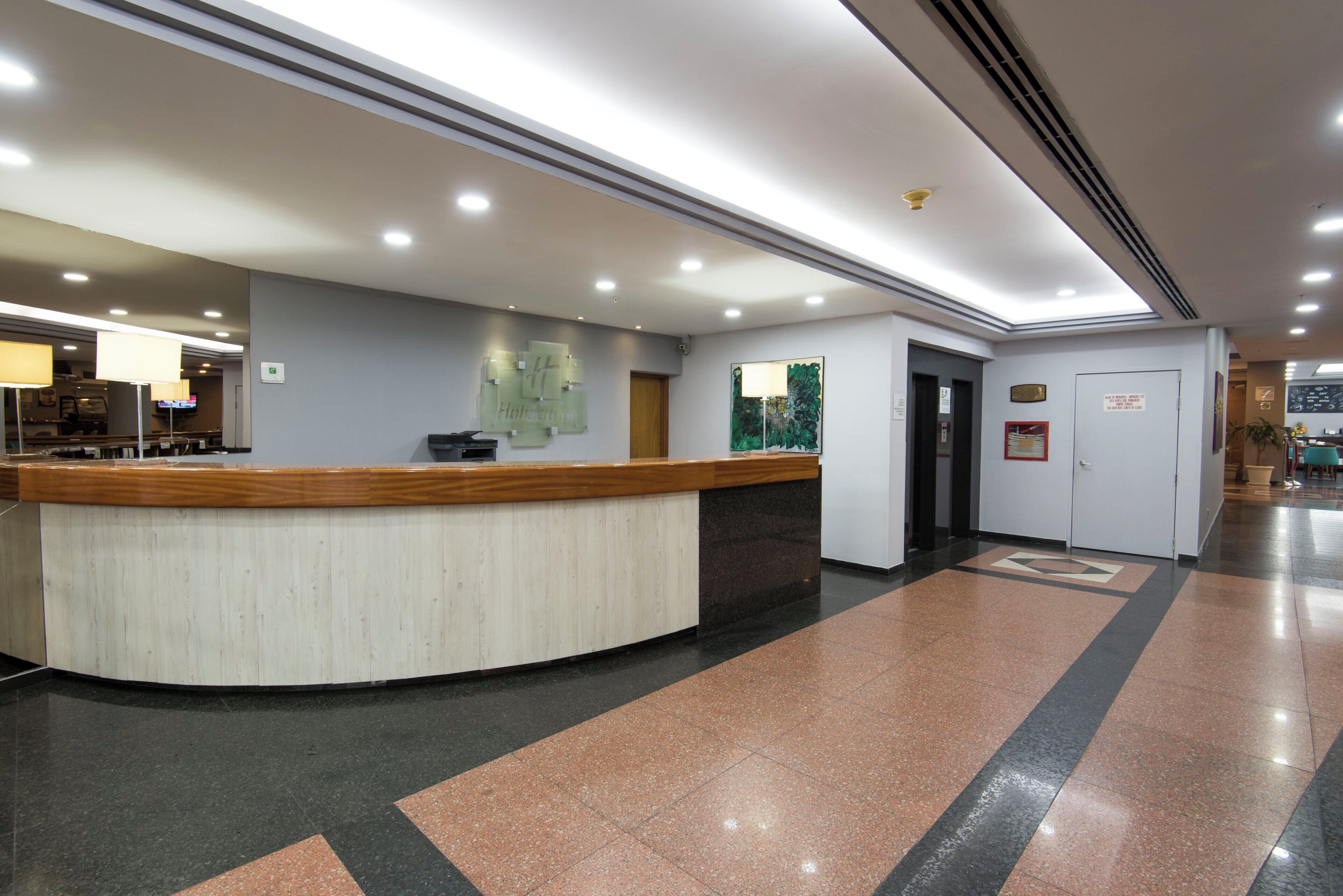 A hotel reception area with a curved reception desk, a wall-mounted information board, and a door leading to another room.