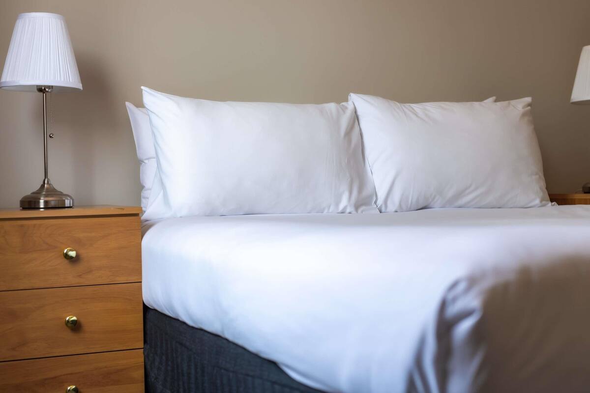 A neatly made bed with white linens and two pillows, a bedside lamp, and a wooden nightstand with drawers.