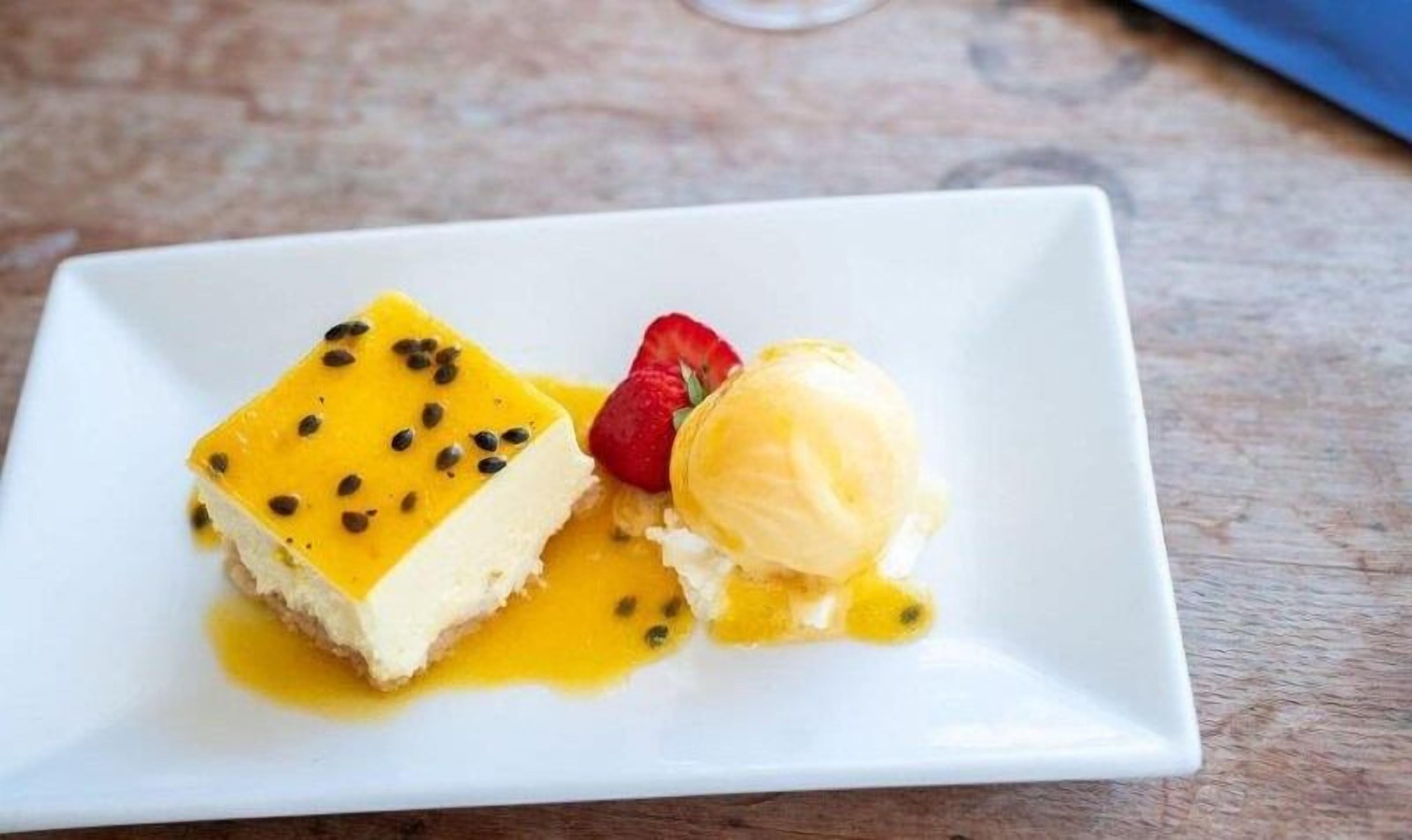 Prato de sobremesa com uma fatia de cheesecake e uma bola de sorvete de baunilha, decorado com morango e sementes.