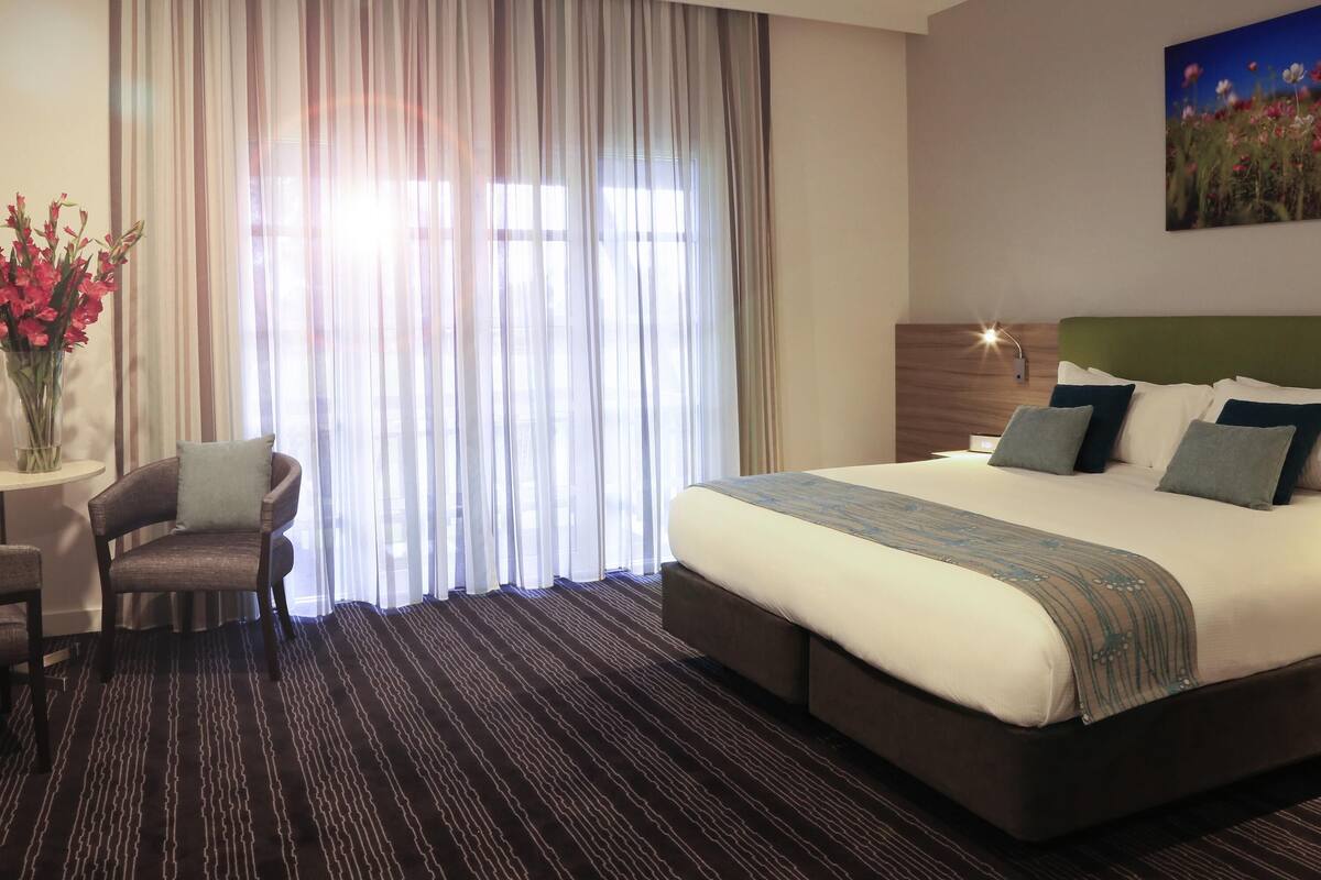 A hotel room with a large bed, a chair, a vase of flowers, and a painting on the wall.