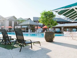 Un área junto a la piscina con sillones de descanso, una jardinera grande con un árbol y sombrillas.