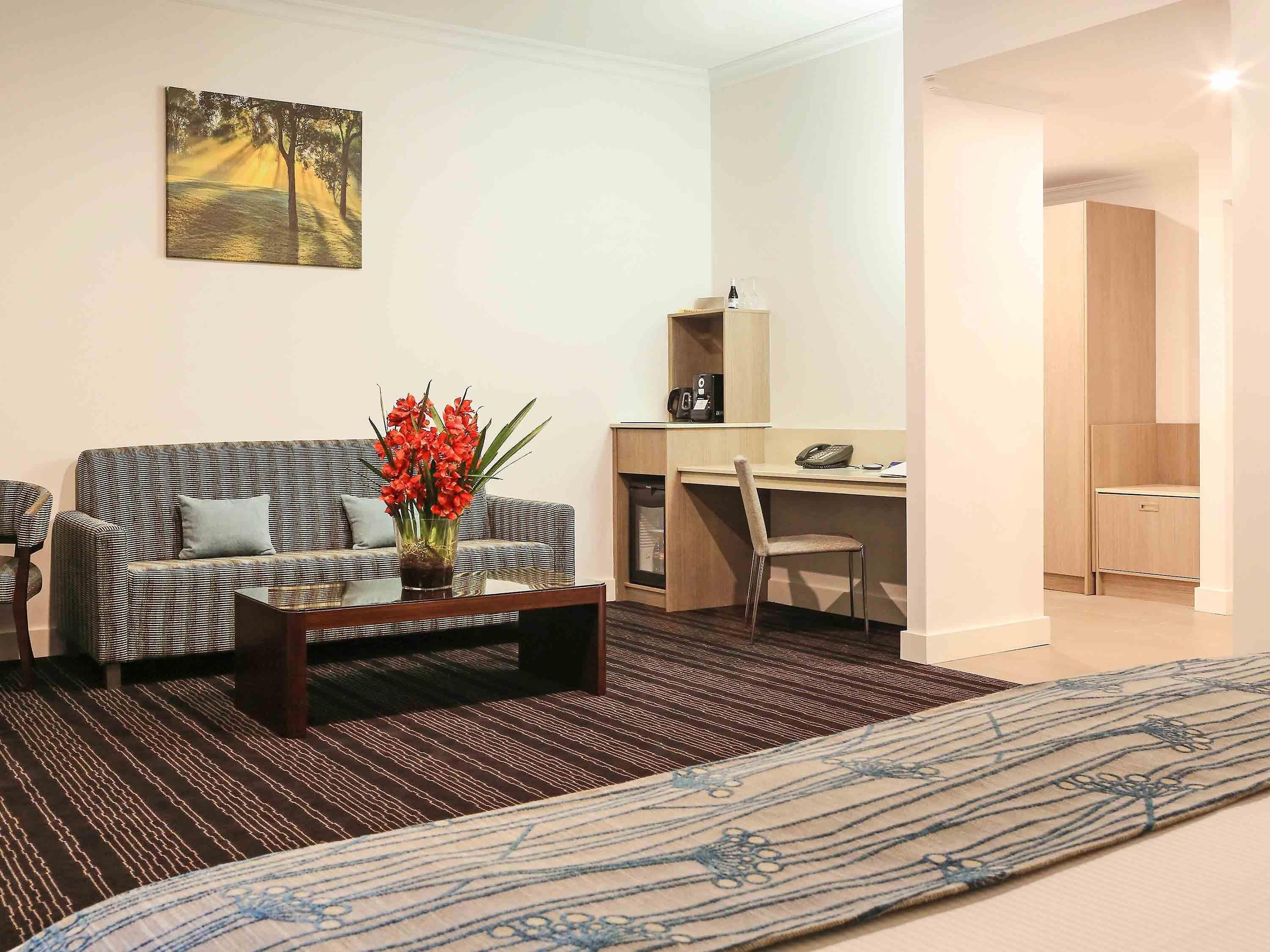 A modern living room with a sofa, a coffee table with a vase of flowers, a desk with a chair, and a bed with a patterned bedspread.