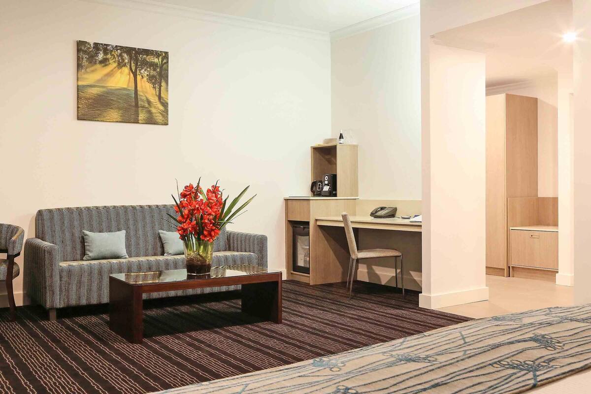 A modern living room with a sofa, a coffee table with a vase of flowers, a desk with a chair, and a bed with a patterned bedspread.