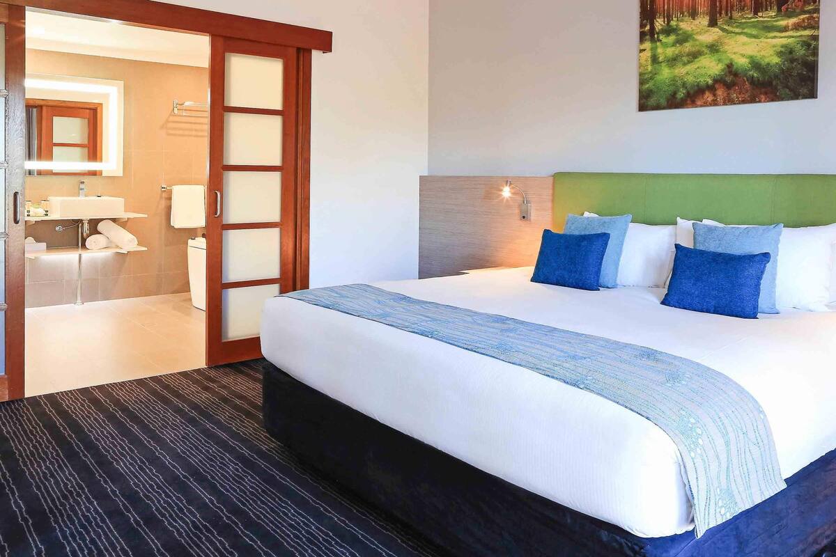 A hotel room with a large bed, a bathroom with a mirror, and a painting on the wall.