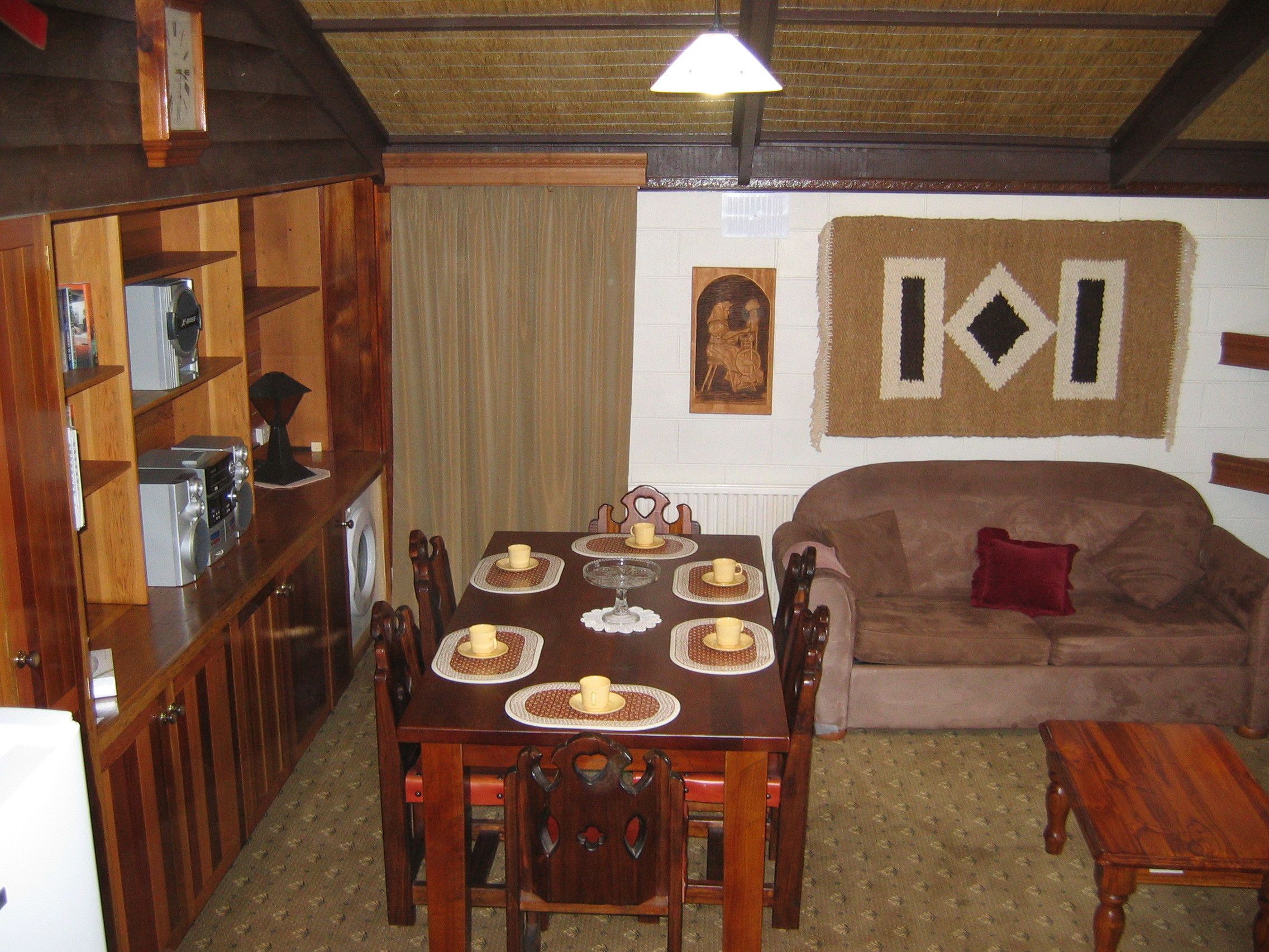 A dining area with a wooden table set for six, surrounded by chairs. A brown sofa and a small wooden table are against the wall.