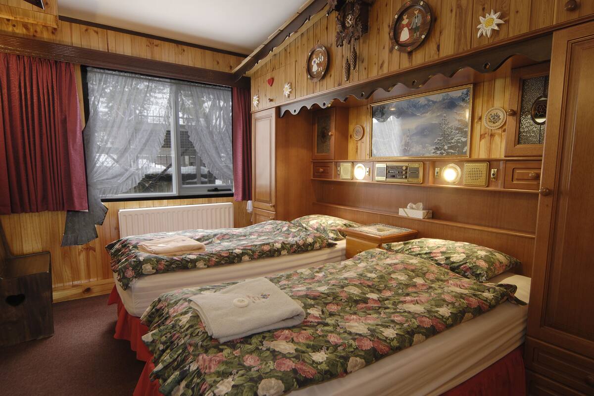 A cabin-style bedroom with two beds, a window with curtains, wooden paneling, and a clock on the wall.