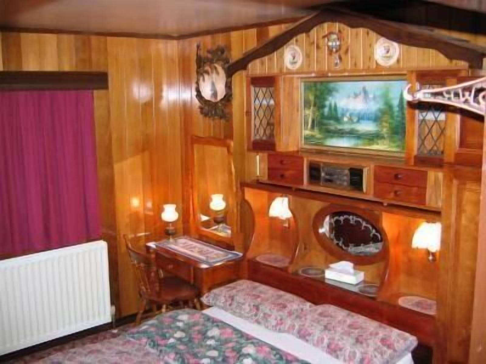 Una habitación de madera con una cama, un escritorio y una silla. Hay dos lámparas en el escritorio y un cuadro en la pared.