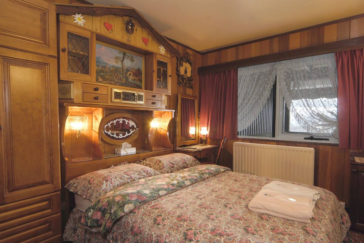 A room with two beds, wooden paneling, a large window with curtains, and a wall-mounted clock.