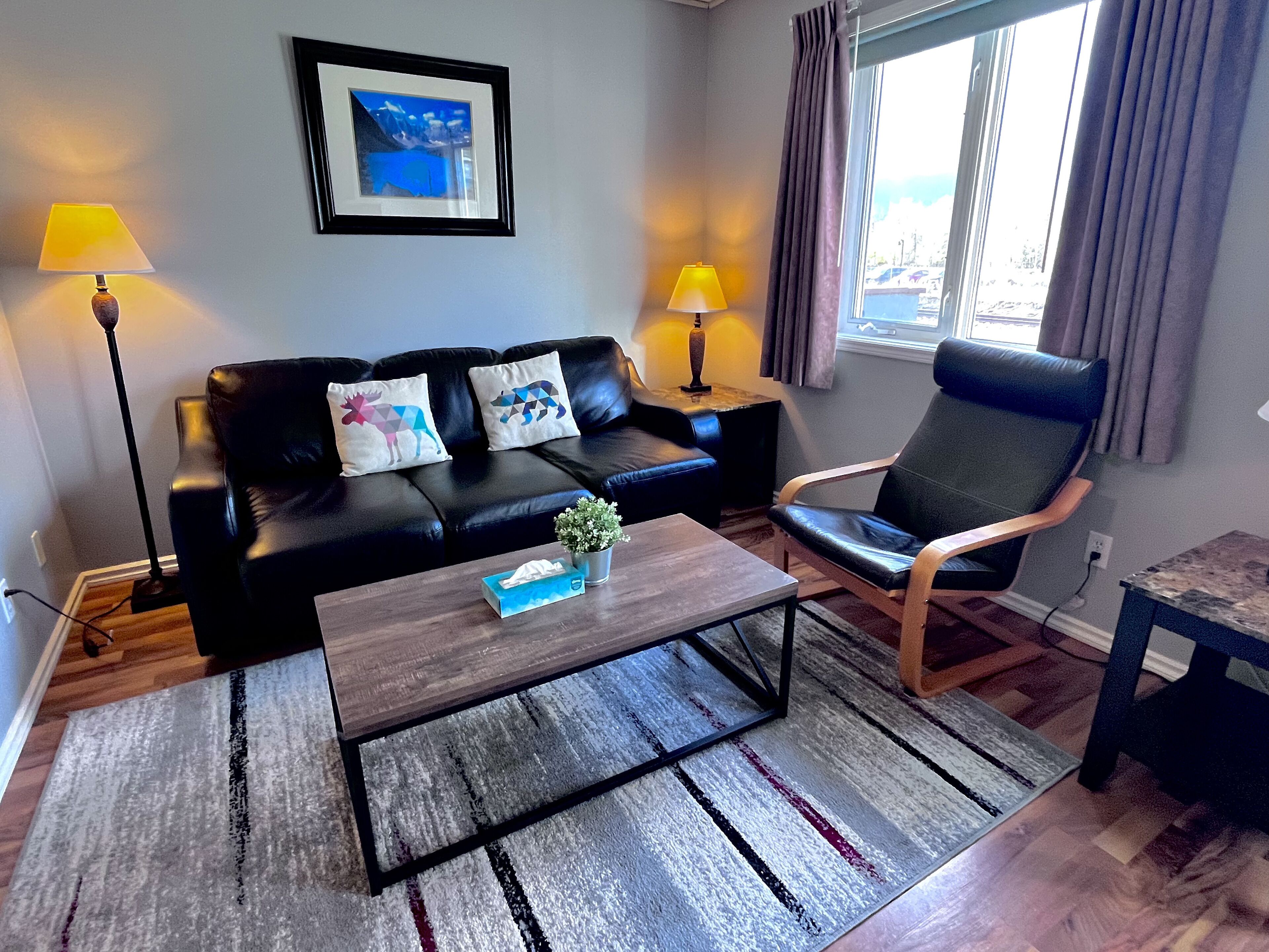 A modern living room with a black leather sofa, two decorative pillows, a wooden coffee table, and a black armchair. There are two floor lamps and a framed picture on the wall.