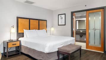 Habitación de hotel con una cama grande, mesitas de noche, un banco, un cuadro enmarcado y un baño con espejo y lavamanos.