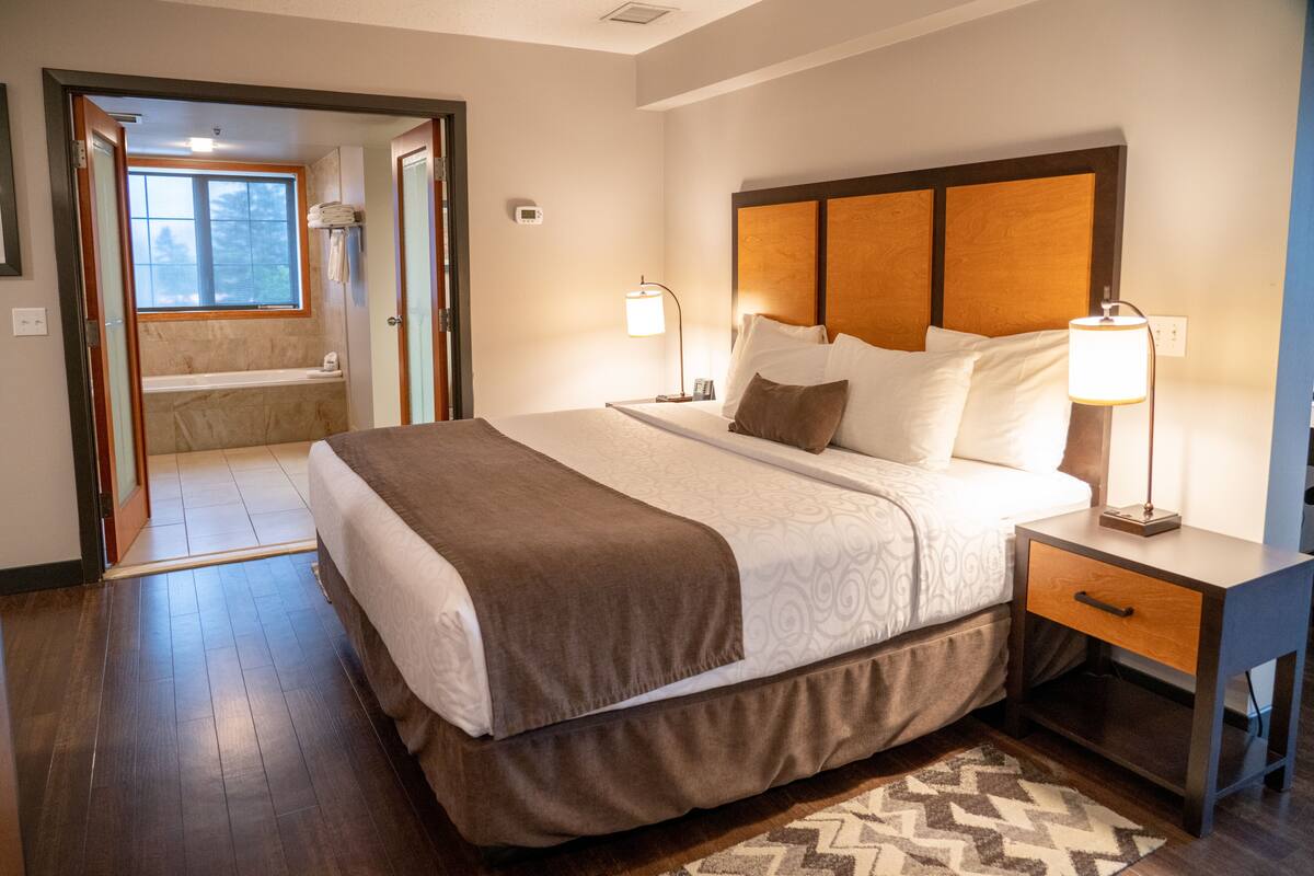 A hotel room with a large bed, bedside lamps, a nightstand, and a view of a bathroom with a bathtub.