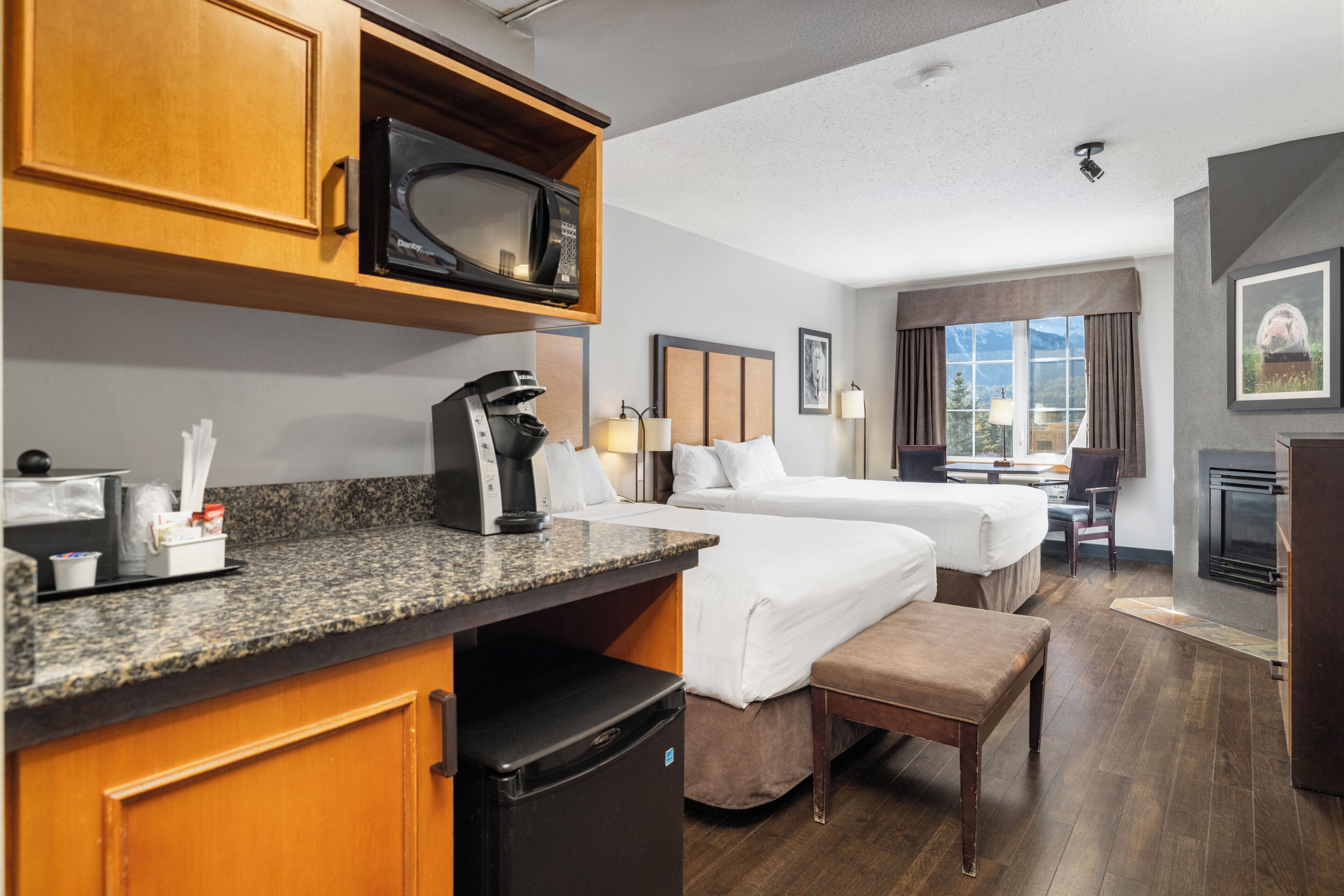 A hotel room with a kitchenette, two beds, a desk, and a view of the outdoors.
