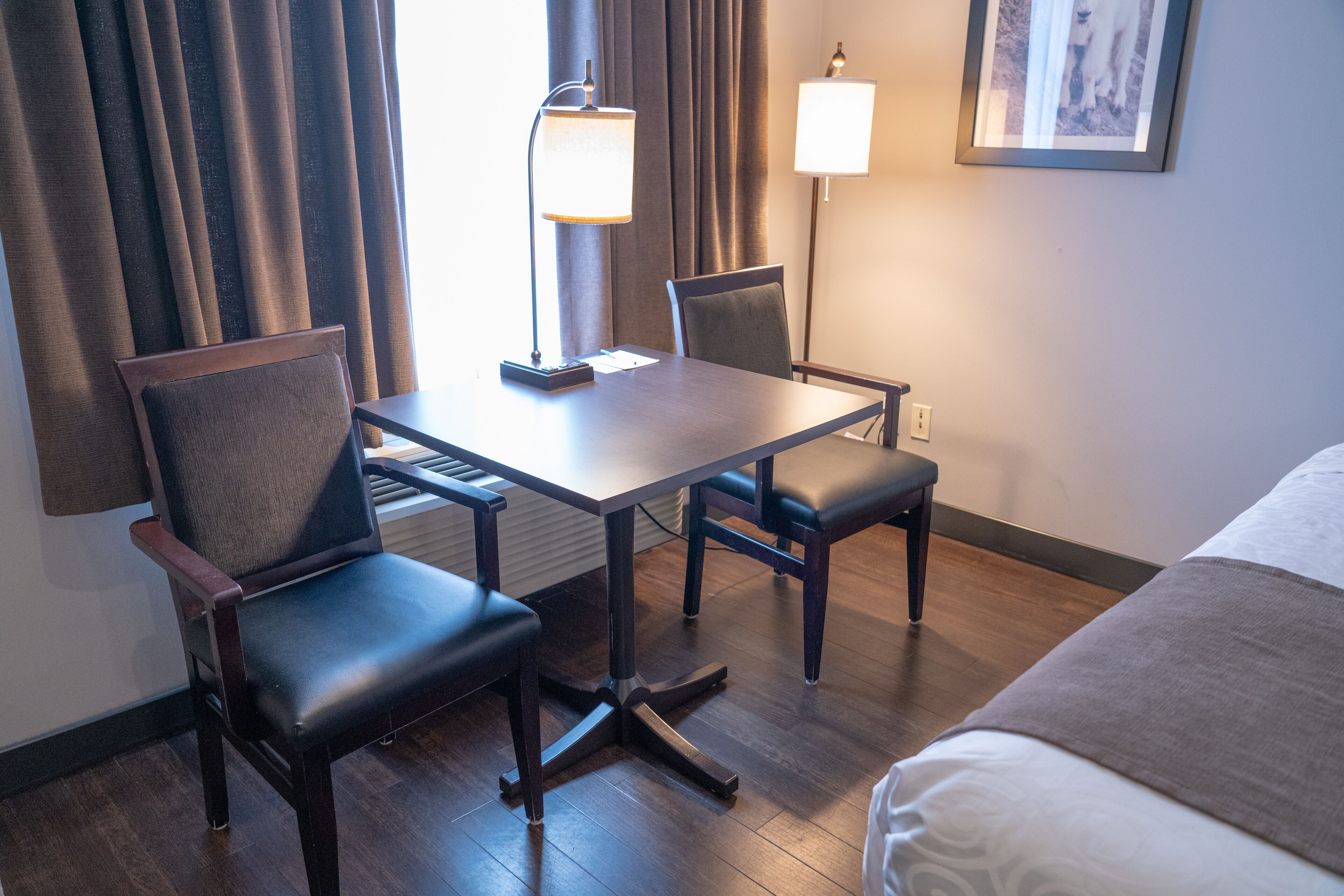 A hotel room with a bed, a desk with two chairs, a lamp, and a framed picture on the wall.