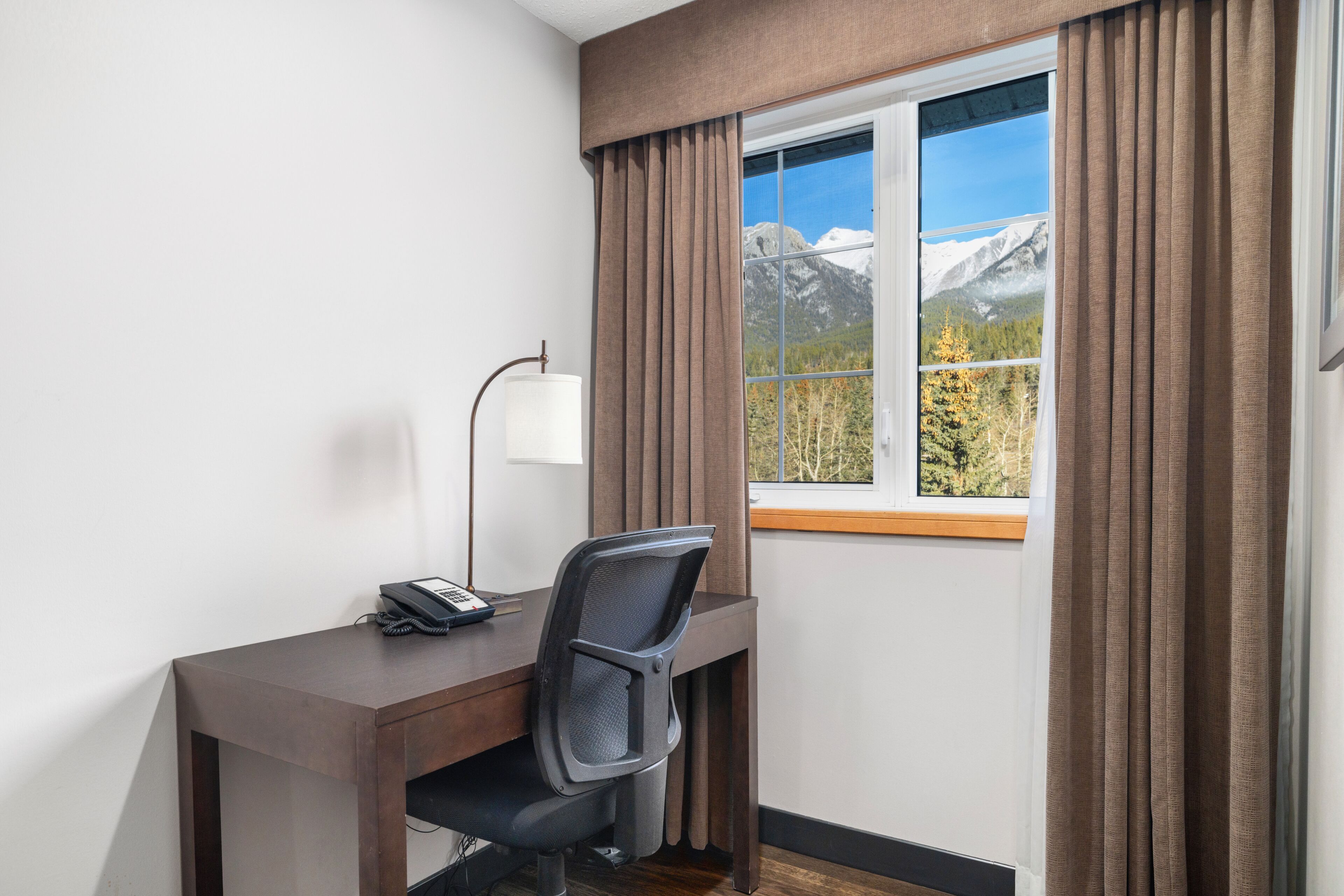 A hotel room with a desk, chair, phone, lamp, and a window with curtains.