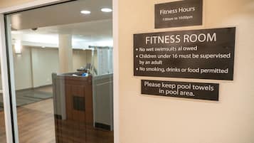 Un gimnasio con un cartel que indica el horario de funcionamiento, de 7:00 a las 22:00, normas sobre trajes de baño mojados, supervisión para menores de 16 años, prohibición de fumar, consumir bebidas o alimentos, y la recomendación de mantener las toallas de piscina en la zona de la piscina.