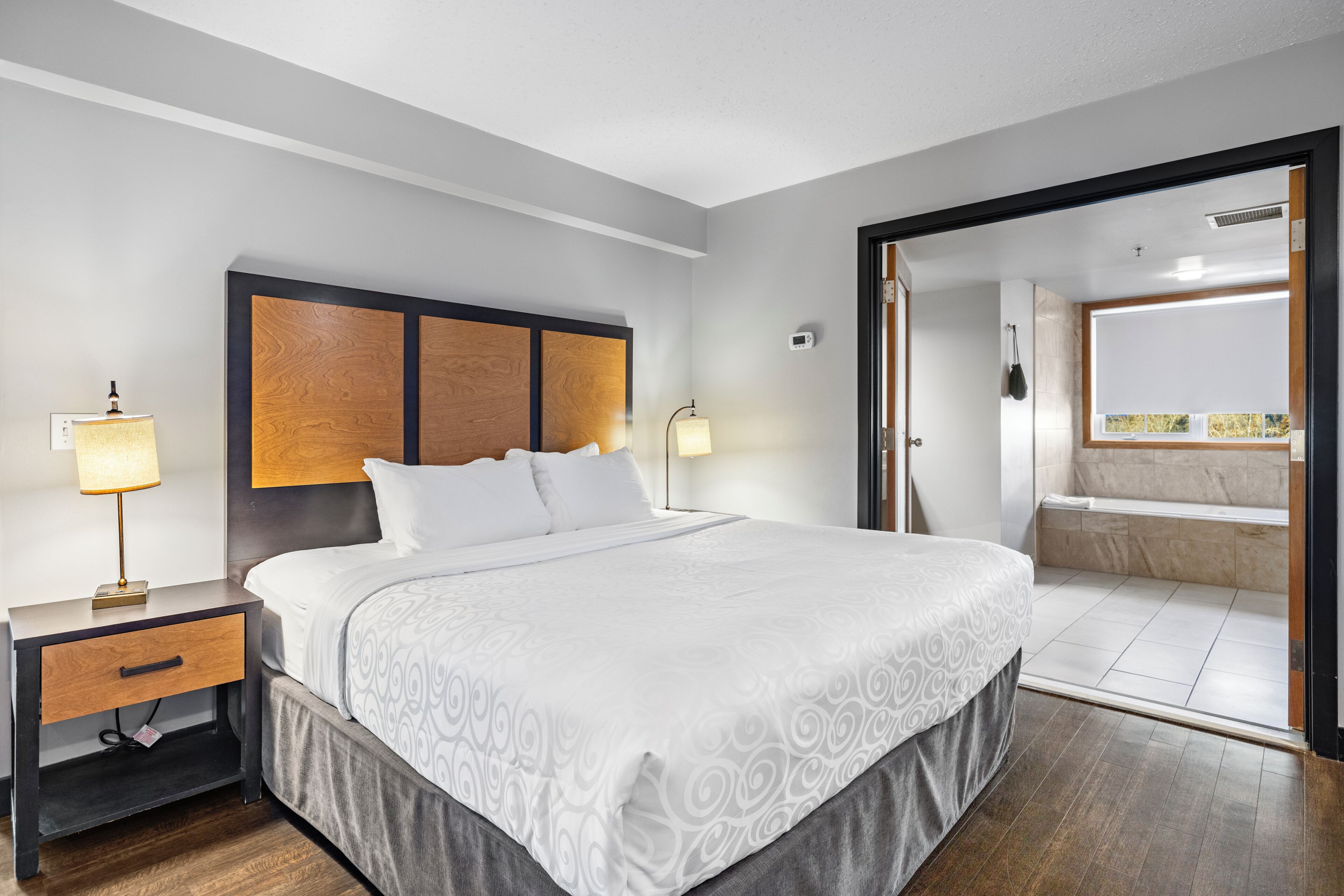 Habitación de hotel con una cama grande, mesitas de noche, una lámpara y vistas al baño.