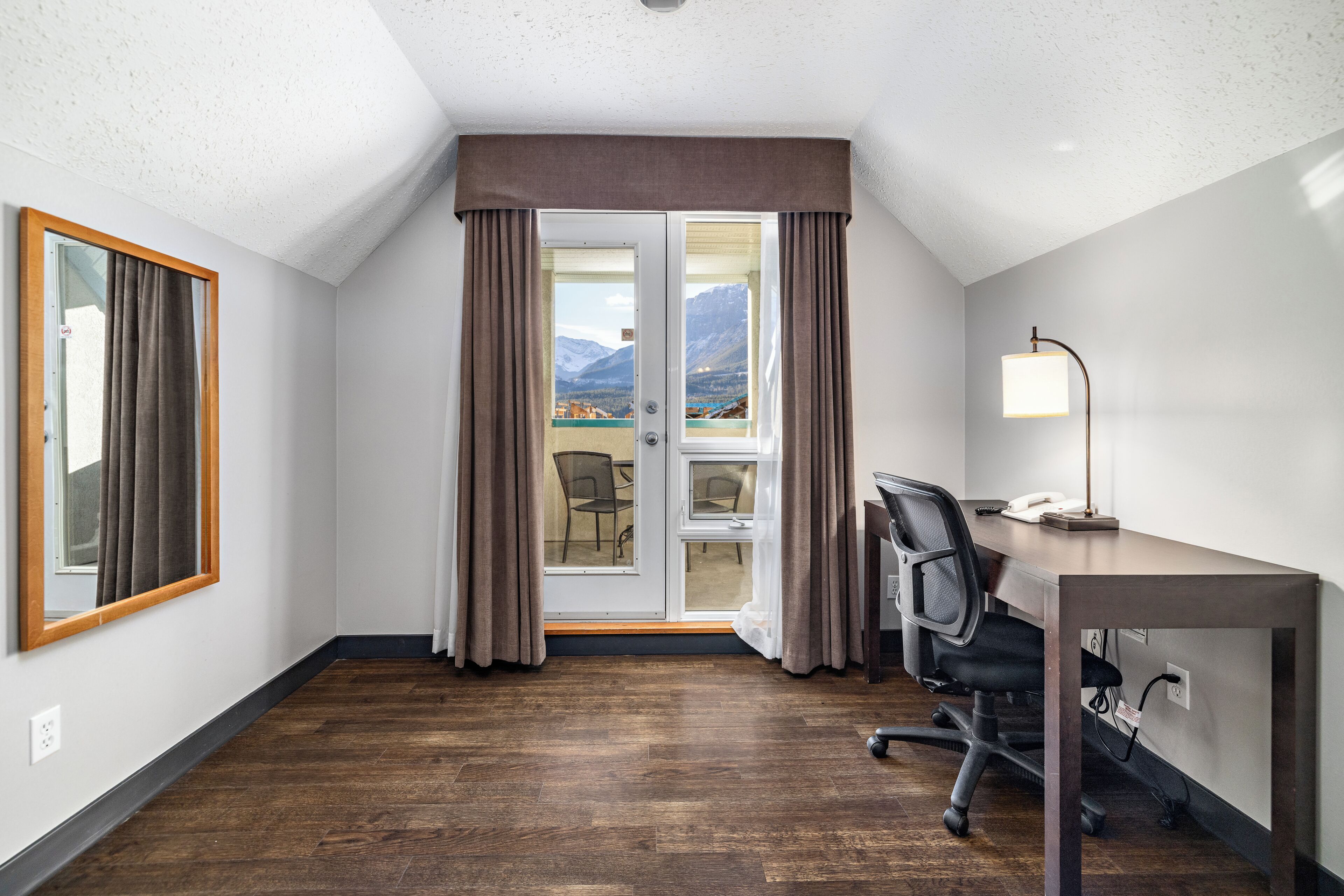 A hotel room with a wooden floor, a desk with a chair, a lamp, and a view of mountains through a glass door.
