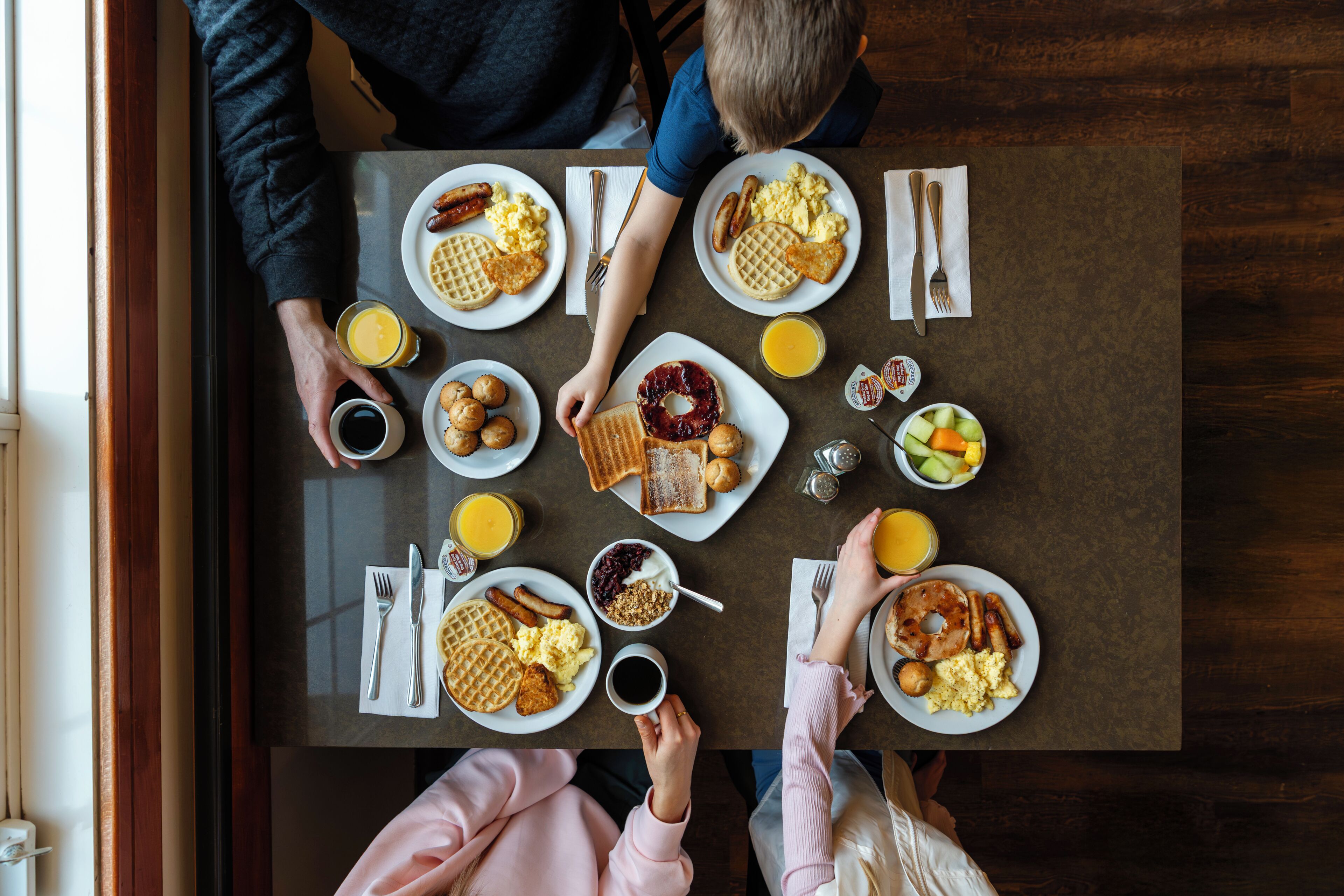 A dining table with breakfast items including waffles, sausages, and toast, with people seated around it.
