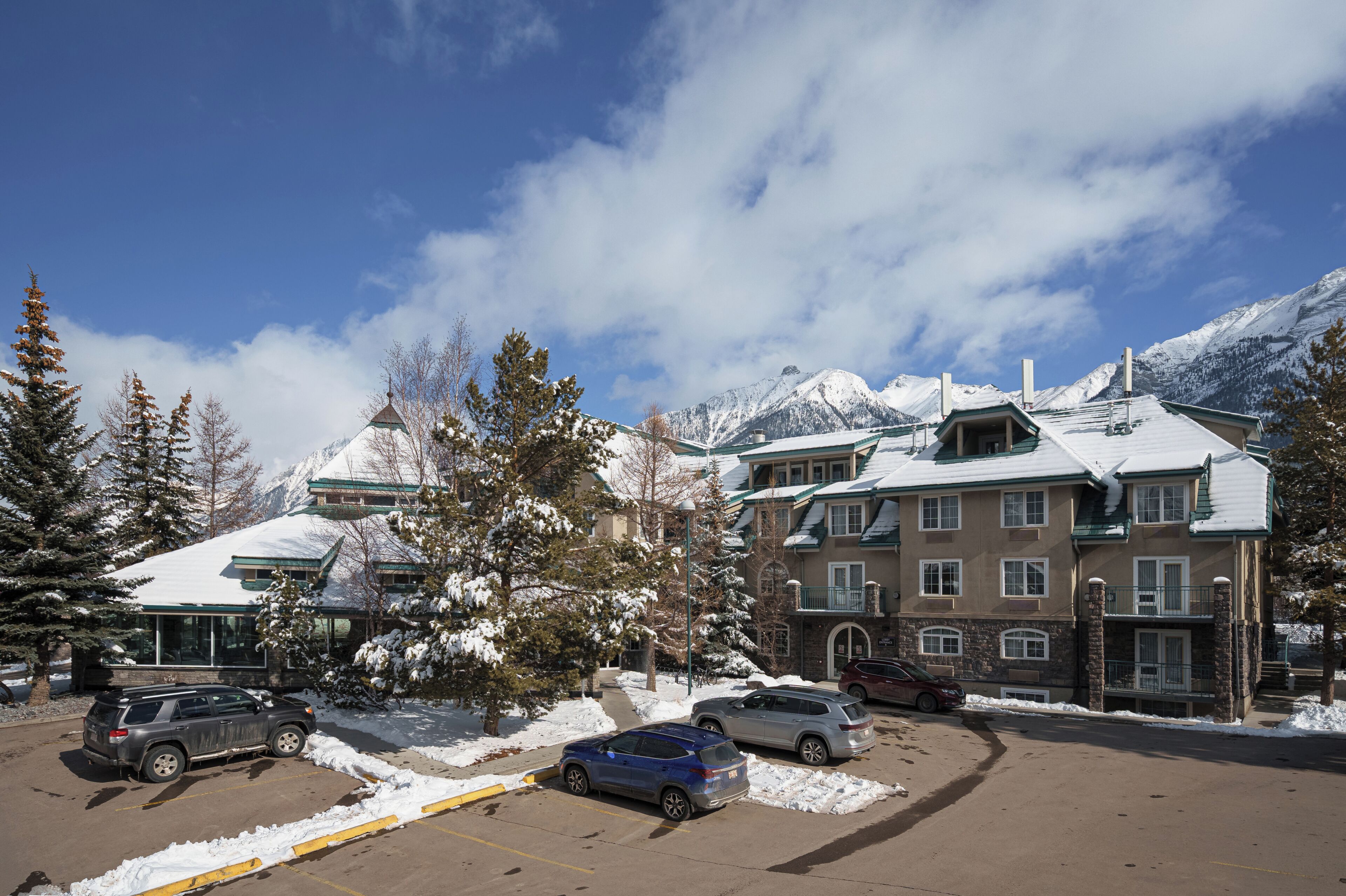 A multi-story hotel with a snow-covered roof, parking lot, and mountains in the background.