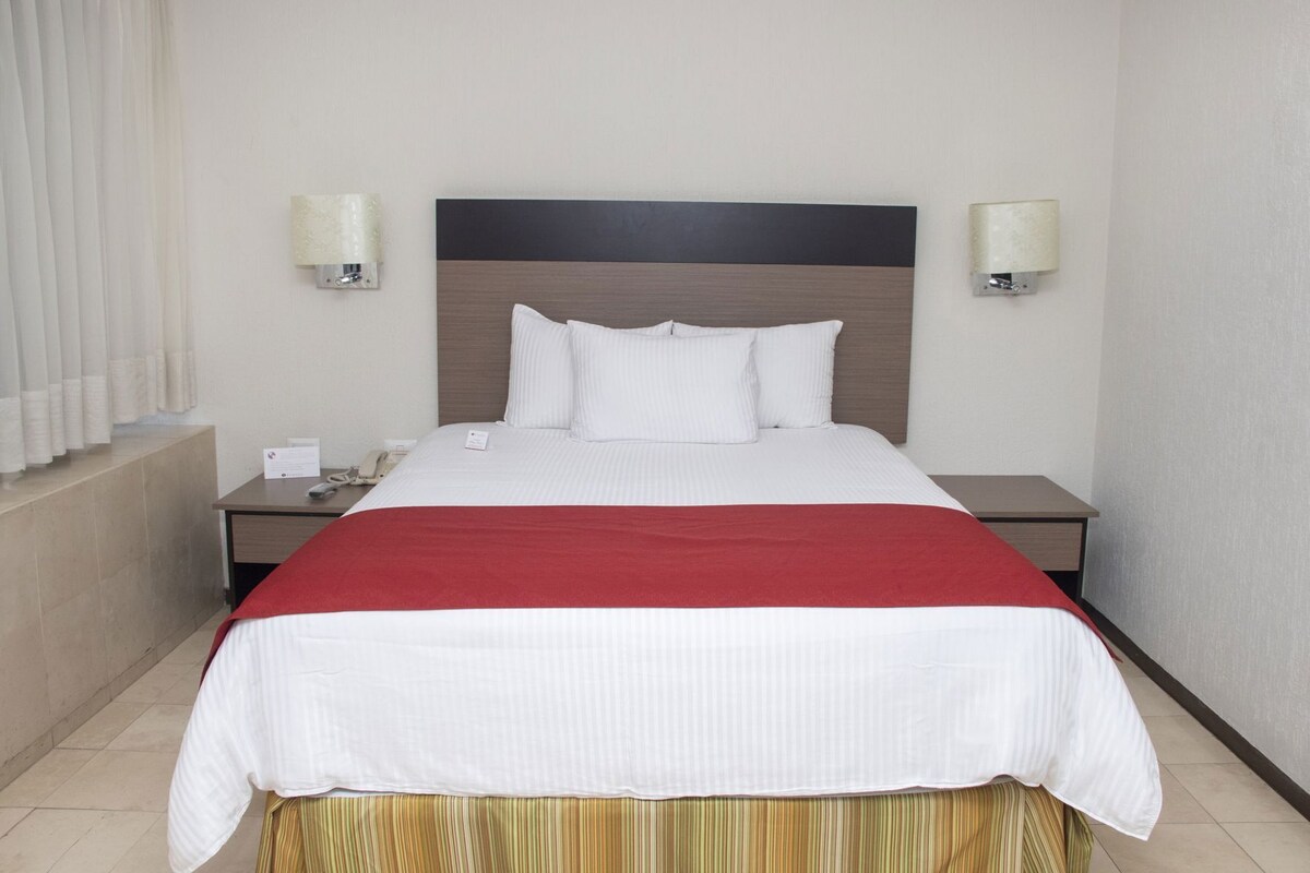 Una habitación de hotel con cama, mesitas de noche, teléfono y lámparas de pared.
