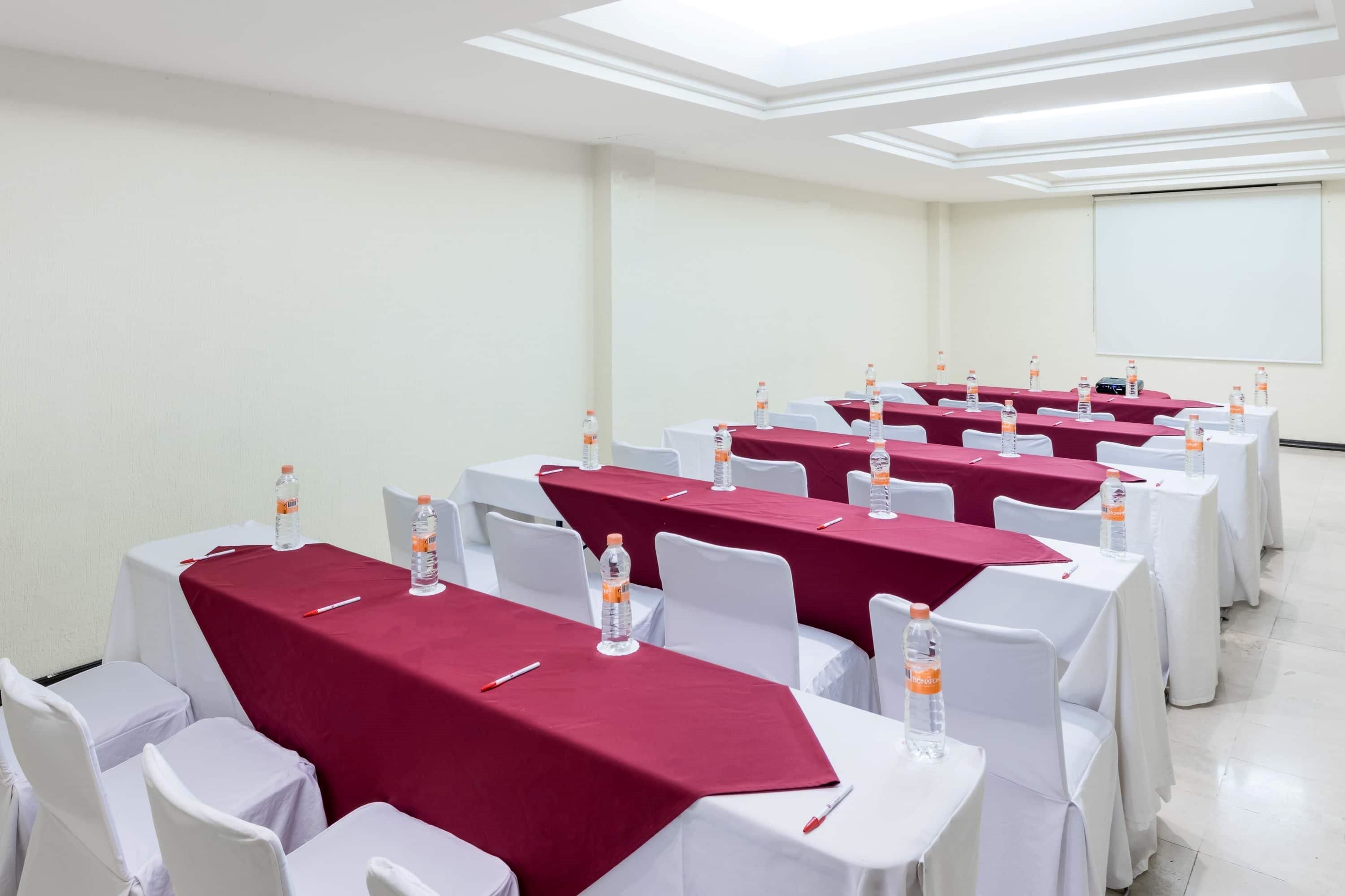 A conference room with rows of tables covered in red tablecloths, white chairs, and water bottles.