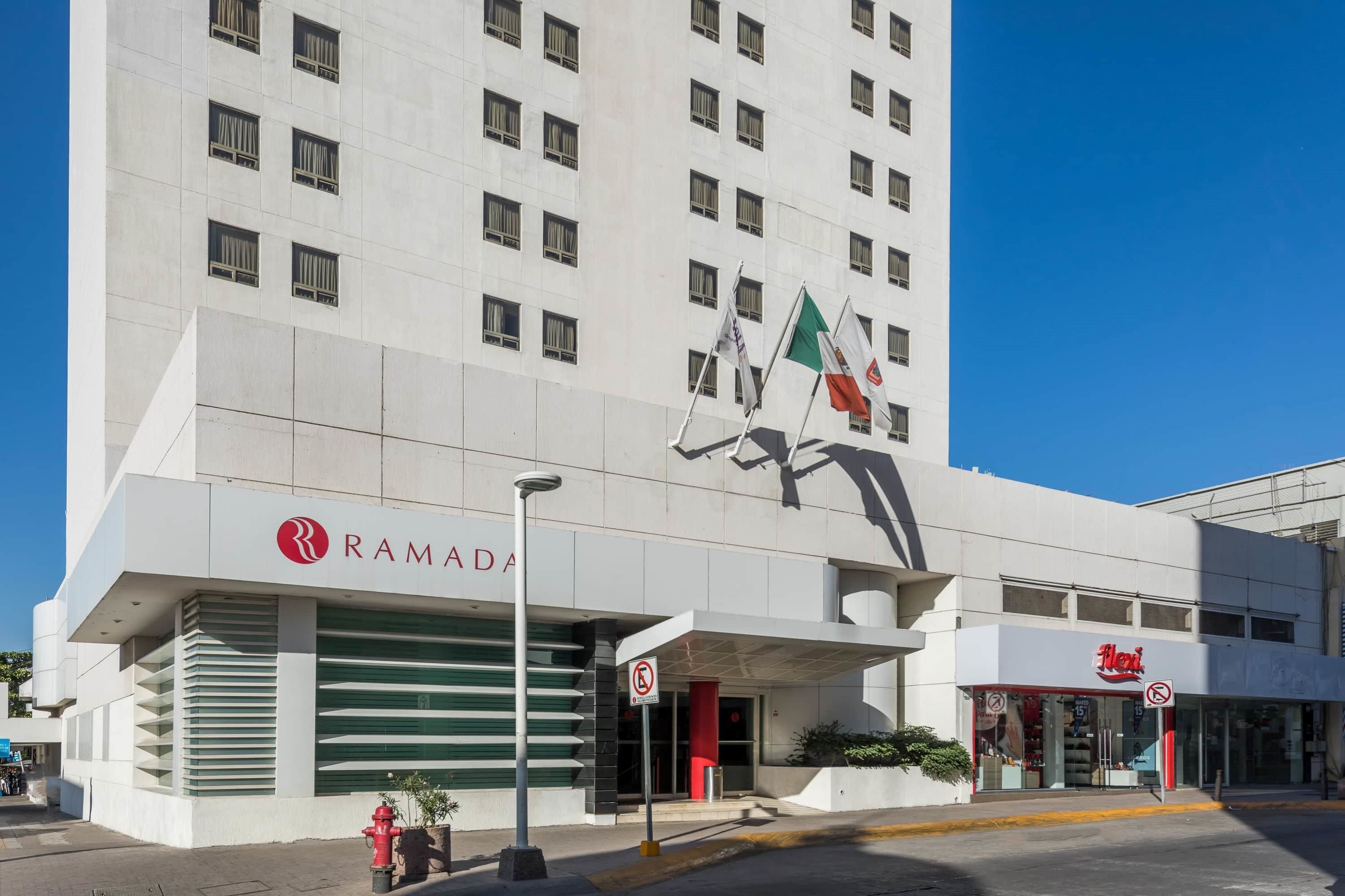 Un hôtel moderne avec un grand logo sur la façade, une entrée couverte et une devanture agrémentée d’une enseigne.