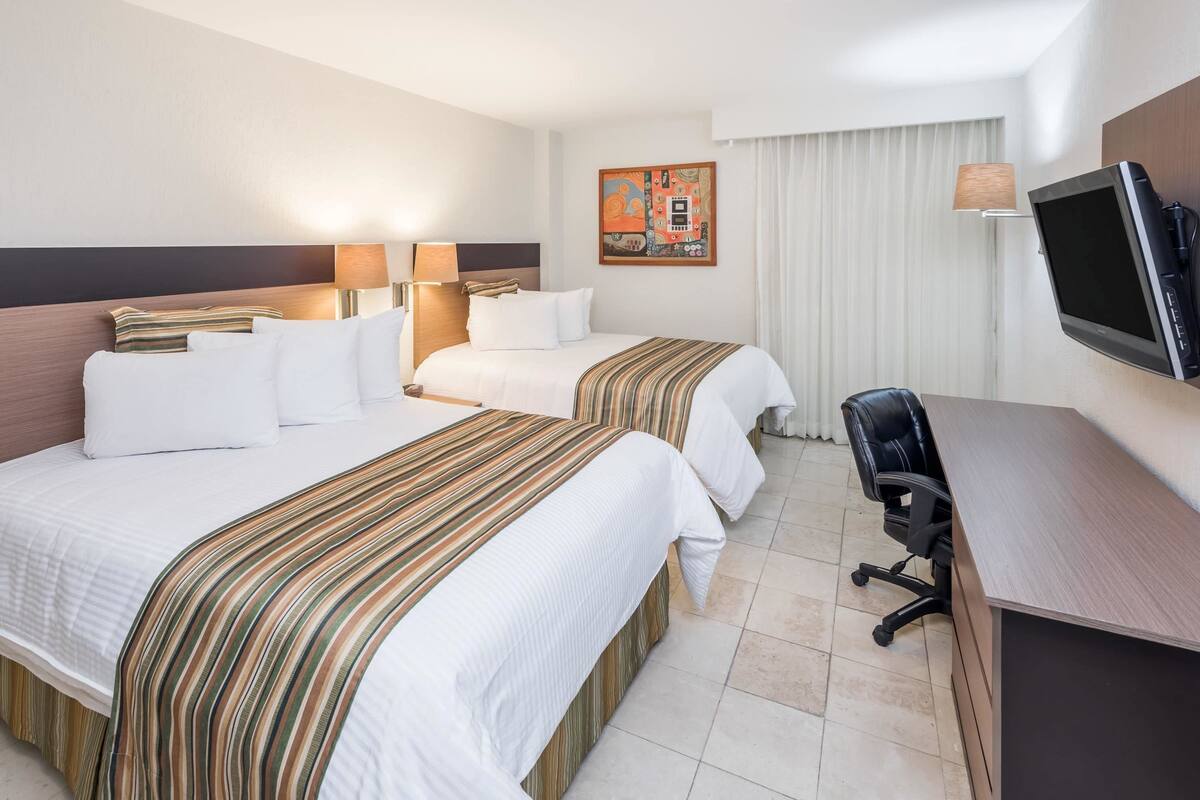 Habitación de hotel con dos camas, un escritorio, una silla, un televisor y un cuadro en la pared.