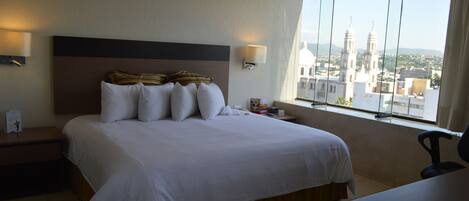 A hotel room with a large bed, a desk, and a window with a cityscape view.