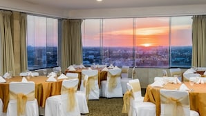 Un salón de banquetes con mesas redondas, sillas blancas con cintas doradas y vistas a la ciudad al atardecer.